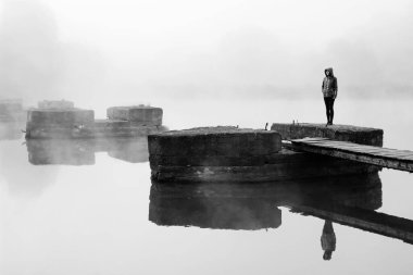 Nehirli mistik bir manzara. Minimalist siyah beyaz fotoğraf. Kız sabah sisli, yıkılmış eski köprüde duruyor.