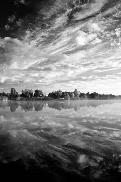 Cloudy vertical landscape. Black and white photo. Calm river in morning fog. Reflection of sky in water