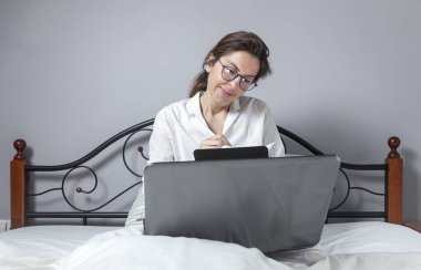 Gözlüklü yetişkin bir kadın, evde pijamaları ve cep telefonuyla çalışıyor. Ev kavramından çalış