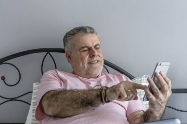 Yatakta pijama, dizüstü bilgisayar ve cep telefonu Coronavirus konseptiyle çalışan sakallı olgun bir adam.