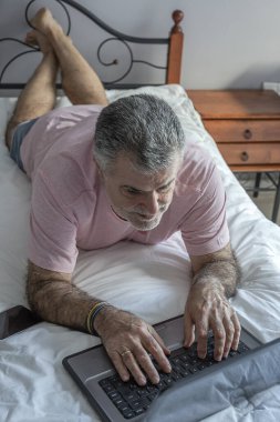 Yatakta pijama, dizüstü bilgisayar ve cep telefonu Coronavirus konseptiyle çalışan sakallı olgun bir adam.