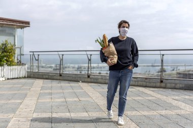 Karantina sırasında koronavirüse karşı koruyucu maskeli olgun bir kadın gerekli ürünleri satın alarak evine dönüyor. Covid-19 konsepti.