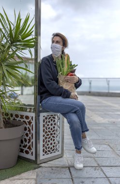 Karantina sırasında koronavirüse karşı koruyucu maskeli olgun bir kadın gerekli ürünleri satın alarak evine dönüyor. Covid-19 konsepti.