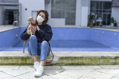 Karantina sırasında koronavirüse karşı koruyucu maskeli bir kadın boş havuzlu bir şehirde köpekle oynuyor. Covid-19 konsepti