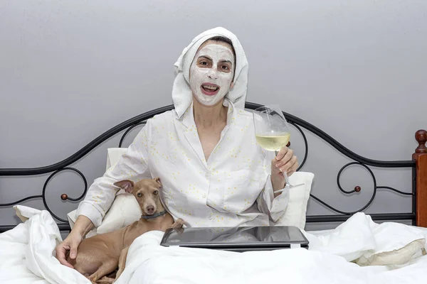 Mature woman taking care of her face with a face mask in bed, with dog, drinking a glass of wine during the covid-19 quarantine.Coronavirus concept