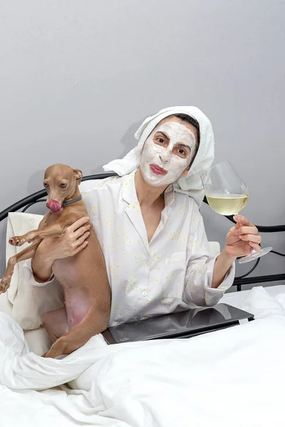 Mature woman taking care of her face with a face mask in bed, with dog, drinking a glass of wine during the covid-19 quarantine.Coronavirus concept