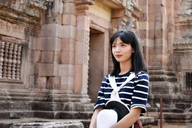 Çizgili gömlek giyen ve elinde beyaz şapkalı genç Asyalı kadın Tayland 'daki Prasat Hin Phanom Rung tapınağının kalıntılarını araştırıyor.