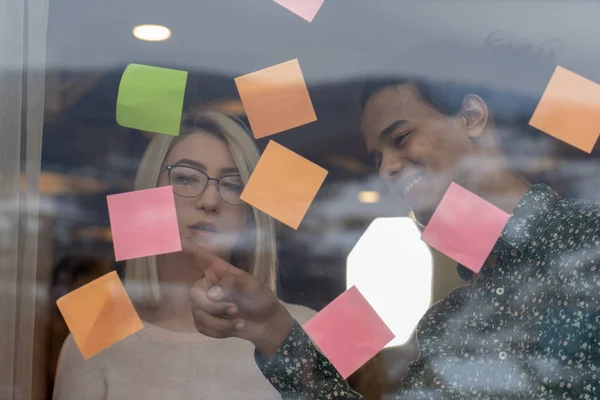 positive young people laughing while working together during brainstorming and standing behind glass wall with sticky colorful papers.Cheerful students learning words from stickers