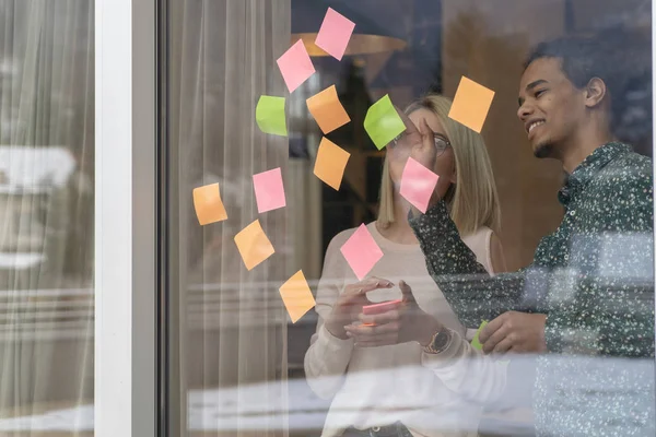 positive young people laughing while working together during brainstorming and standing behind glass wall with sticky colorful papers.Cheerful students learning words from stickers