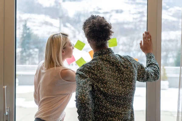 positive young people laughing while working together during brainstorming and standing behind glass wall with sticky colorful papers.Cheerful students learning words from stickers