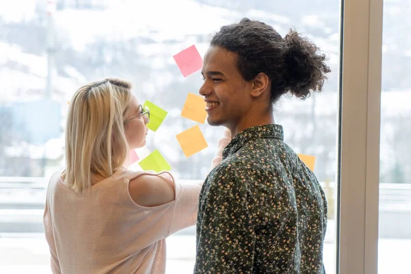 positive young people laughing while working together during brainstorming and standing behind glass wall with sticky colorful papers.Cheerful students learning words from stickers