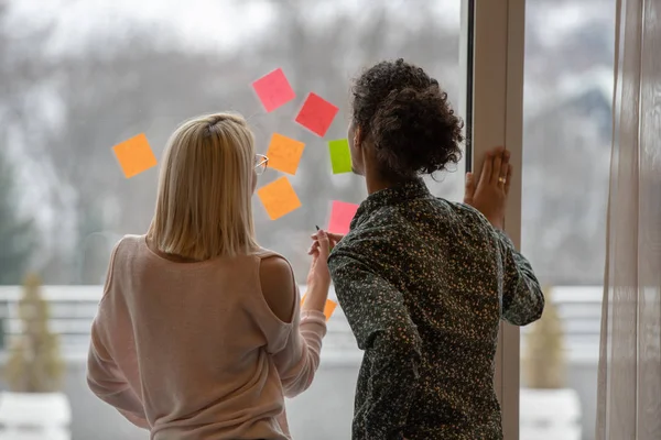 positive young people laughing while working together during brainstorming and standing behind glass wall with sticky colorful papers.Cheerful students learning words from stickers