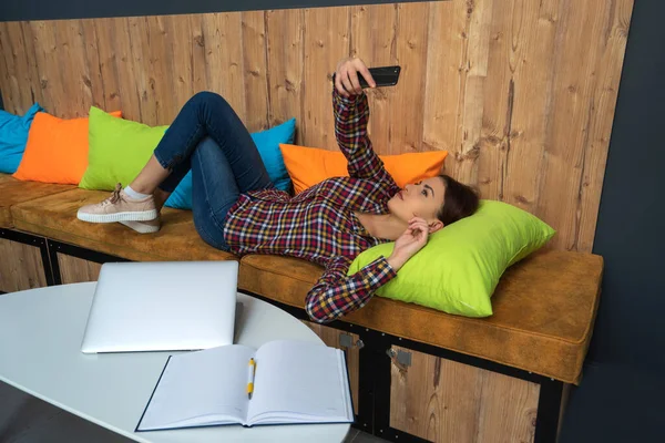 Όμορφη κοπέλα σε casual ρούχα κάνει selfie χρησιμοποιώντας ένα έξυπνο τηλέφωνο και χαμογελώντας, ενώ εργάζονται με ένα φορητό υπολογιστή στο γραφείο. Έννοια εκκίνησης — Φωτογραφία Αρχείου
