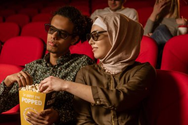 Afrikalı Amerikalı Müslüman adam, karısıyla sinemada oturuyor, 3 boyutlu film izliyor, patlamış mısır yiyor, gülümsüyor..