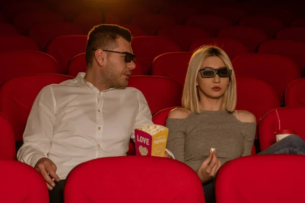 Happy couple sitting in movie theater, watching 3D movie, eating popcorn, smiling
