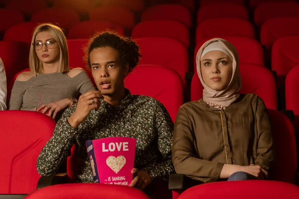 African american muslim man with his wife sitting in movie theater, watching movie, eating popcorn, smiling.