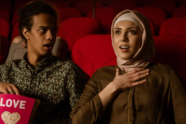 African american muslim man with his wife sitting in movie theater, watching movie, eating popcorn, smiling.