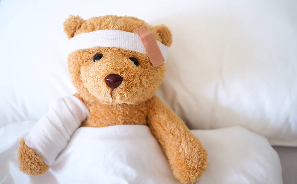 Медведь Тедди лежит больной в постели с повязкой на голове и покрытой тканью.