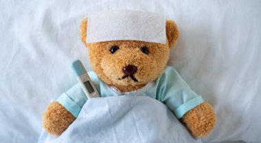The teddy bear is sick on the bed with a high fever. There is a fever reducing sheet on the forehead. clipart
