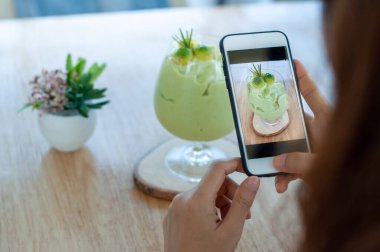 Kahve dükkanında cep telefonu kullanarak ön taraftaki avokado smoothie 'nin fotoğraflarını sosyal medyaya yükleyen bir kadın. Sağlıklı günler.