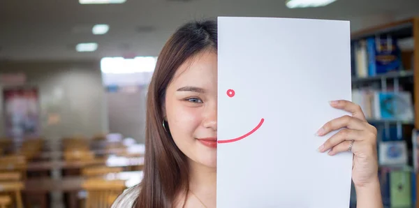 A beautiful female student in Asia holds a paper drawing a smile with red ink and a happy posture on campus.