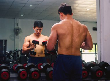 An Asian man lifts a dumbbell in front of a mirror in a gym. Exe