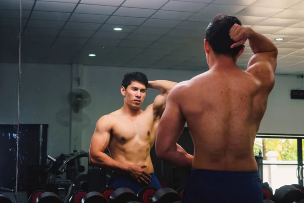 Asiatische junge Mann lässt seine Muskeln vor einem Glas in der g — Stockfoto