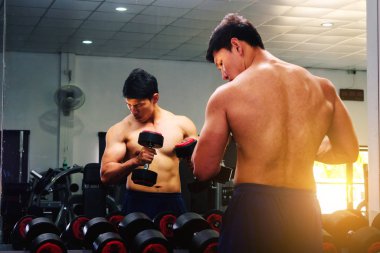An Asian man lifts a dumbbell in front of a mirror in a gym. Exe