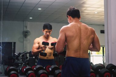 An Asian man lifts a dumbbell in front of a mirror in a gym. Exe