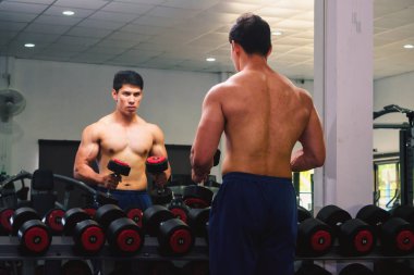 An Asian man lifts a dumbbell in front of a mirror in a gym. Exe