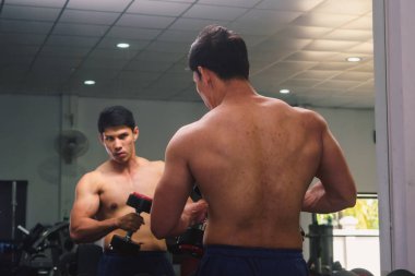 An Asian man lifts a dumbbell in front of a mirror in a gym. Exe