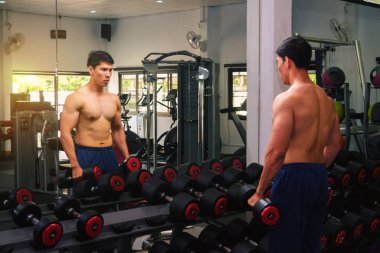 An Asian man lifts a dumbbell in front of a mirror in a gym. Exe