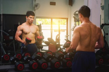An Asian man lifts a dumbbell in front of a mirror in a gym. Exe