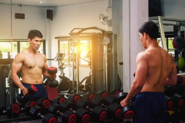 An Asian man lifts a dumbbell in front of a mirror in a gym. Exe