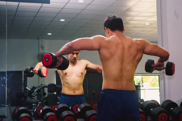 An Asian man lifts a dumbbell in front of a mirror in a gym. Exe — ストック写真