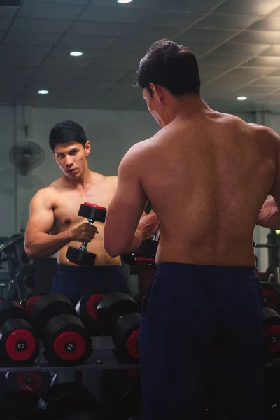 An Asian man lifts a dumbbell in front of a mirror in a gym. Exe