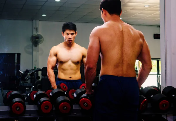 An Asian man lifts a dumbbell in front of a mirror in a gym. Exe — ストック写真