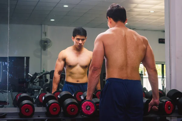 An Asian man lifts a dumbbell in front of a mirror in a gym. Exe — ストック写真