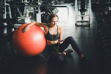 Spor gömlek giyen güzel bir kadın kulaklıklardan müzik dinleyerek rahatlıyor. Lastik yoga topu ya da egzersiz topuyla.