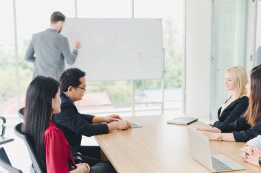 Genç iş adamları beyaz tahtadaki grafikleri ve istatistikleri açıklıyorlar. Toplantıya katılan şirket yöneticileri ve çalışanları.