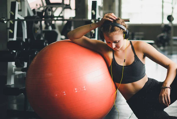 Spor gömlek giyen güzel bir kadın kulaklıklardan müzik dinleyerek rahatlıyor. Lastik yoga topu ya da egzersiz topuyla.