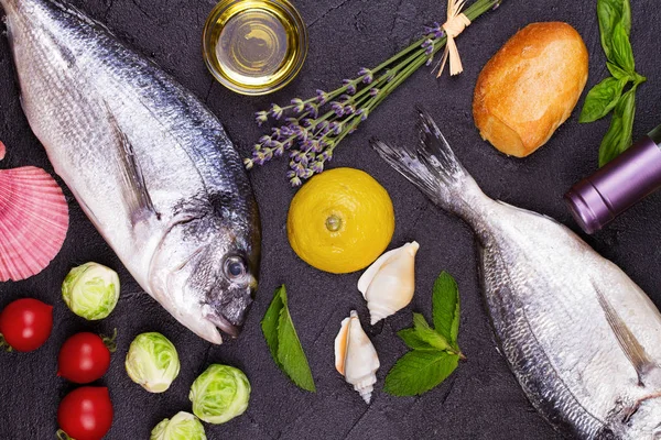 Raw fresh dorado fish with brussels sprouts, tomatoes, lemon, young potato, greens, bread, white wine bottle and olive oil on dark background — Stock Photo, Image