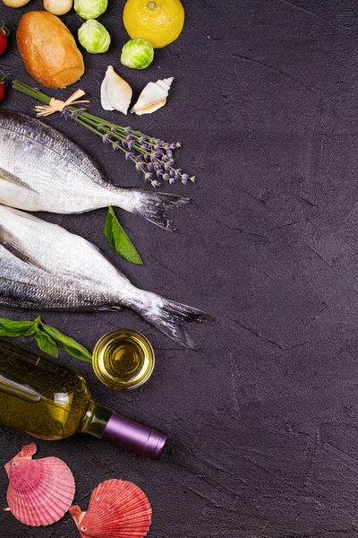 Сырая свежая рыба Дорадо с брюссельской капустой, помидорами, лимоном, картофелем, зеленью, хлебом, бутылкой белого вина и оливковым маслом на тёмном фоне — стоковое фото