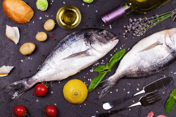 Raw fresh dorado fish with brussels sprouts, tomatoes, lemon, young potato, greens, bread, white wine bottle and olive oil on dark background — Stock Photo, Image