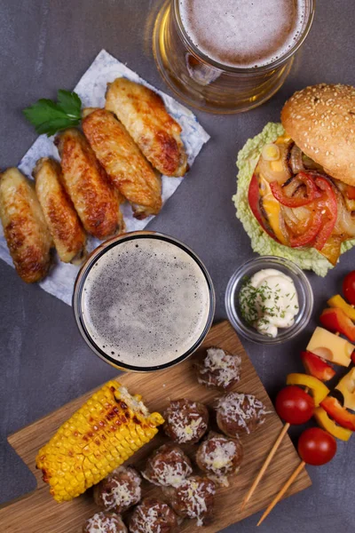 Glasses of beer with chicken wings, burger, meat balls, grilled corn and vegetables
