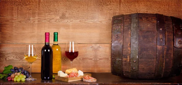 Still life with bottles and glasses of red and white wine with cheese, prosciutto and grape in wine cellar, old wine barrel — Stock Photo, Image