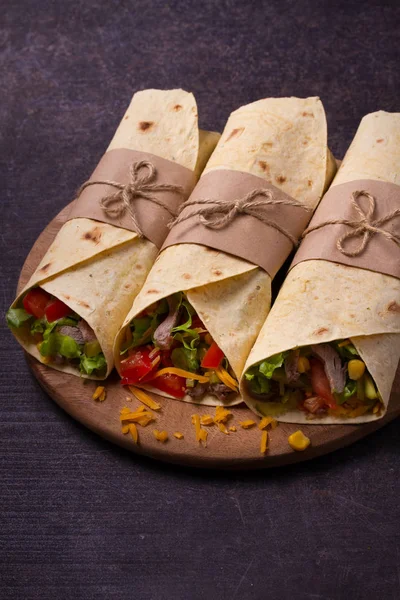 Wraps with beef, tomatoes, lettuce, cucumbers, cheese and sweet corn on cutting board. Tortilla, burritos, sandwiches twisted rolls.