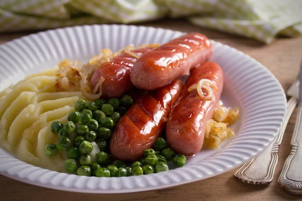 Bangers and Mash. Sausages, mashed potato, green peas and fried onion on white plate on wooden table. horizontal