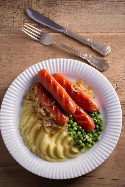 Bangers and Mash. Sausages, mashed potato, green peas and fried onion on white plate on wooden table. overhead, vertical