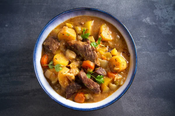 Beef meat stewed with potatoes, carrots and spices in bowl on dark gray background. Overhead horizontal image